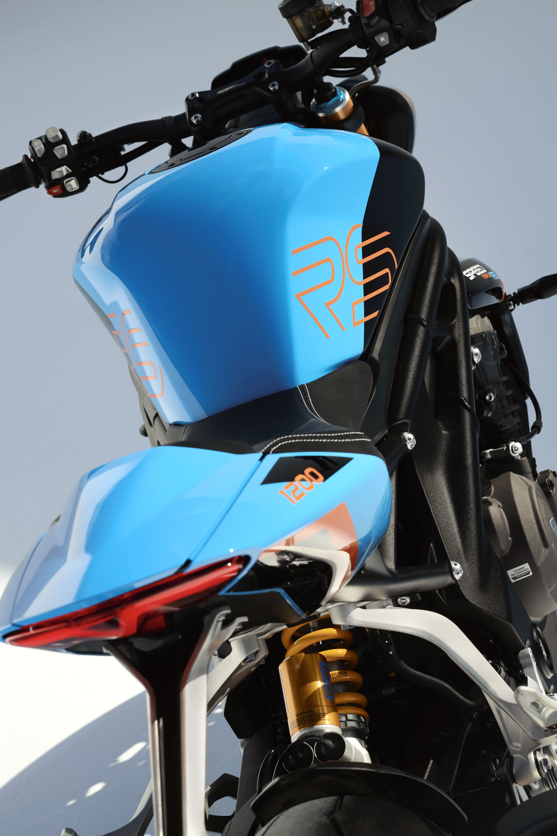A close-up view from behind the fuel tank of a blue and black Triumph Speed Triple 1200 RS, showcasing orange RS lettering, a sharp rear tail section, and a gold Öhlins rear shock absorber.