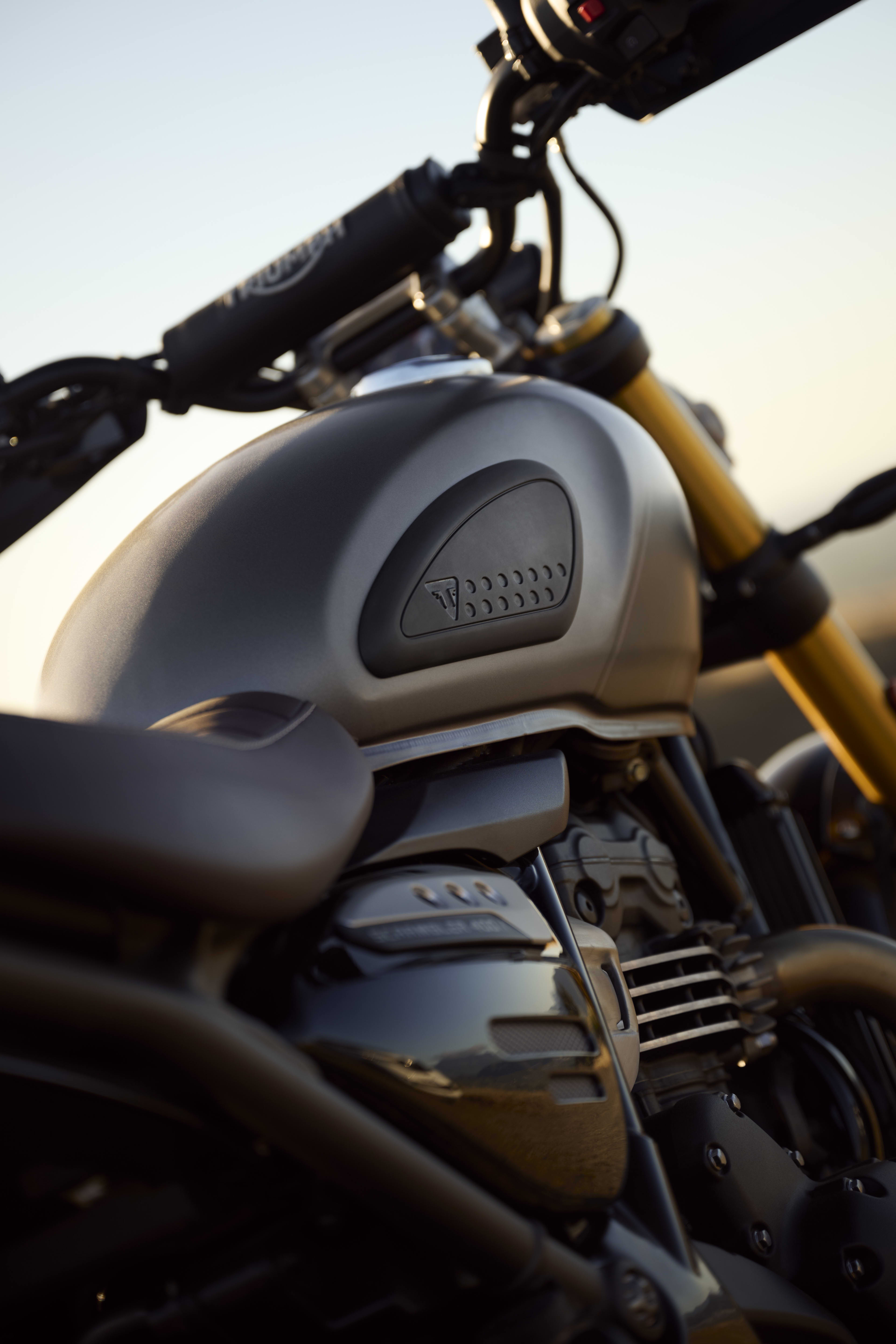 A close-up view of the matte grey fuel tank on a Triumph Scrambler 400 X, featuring black knee pads, gold front forks, and a black handlebar brace pad.