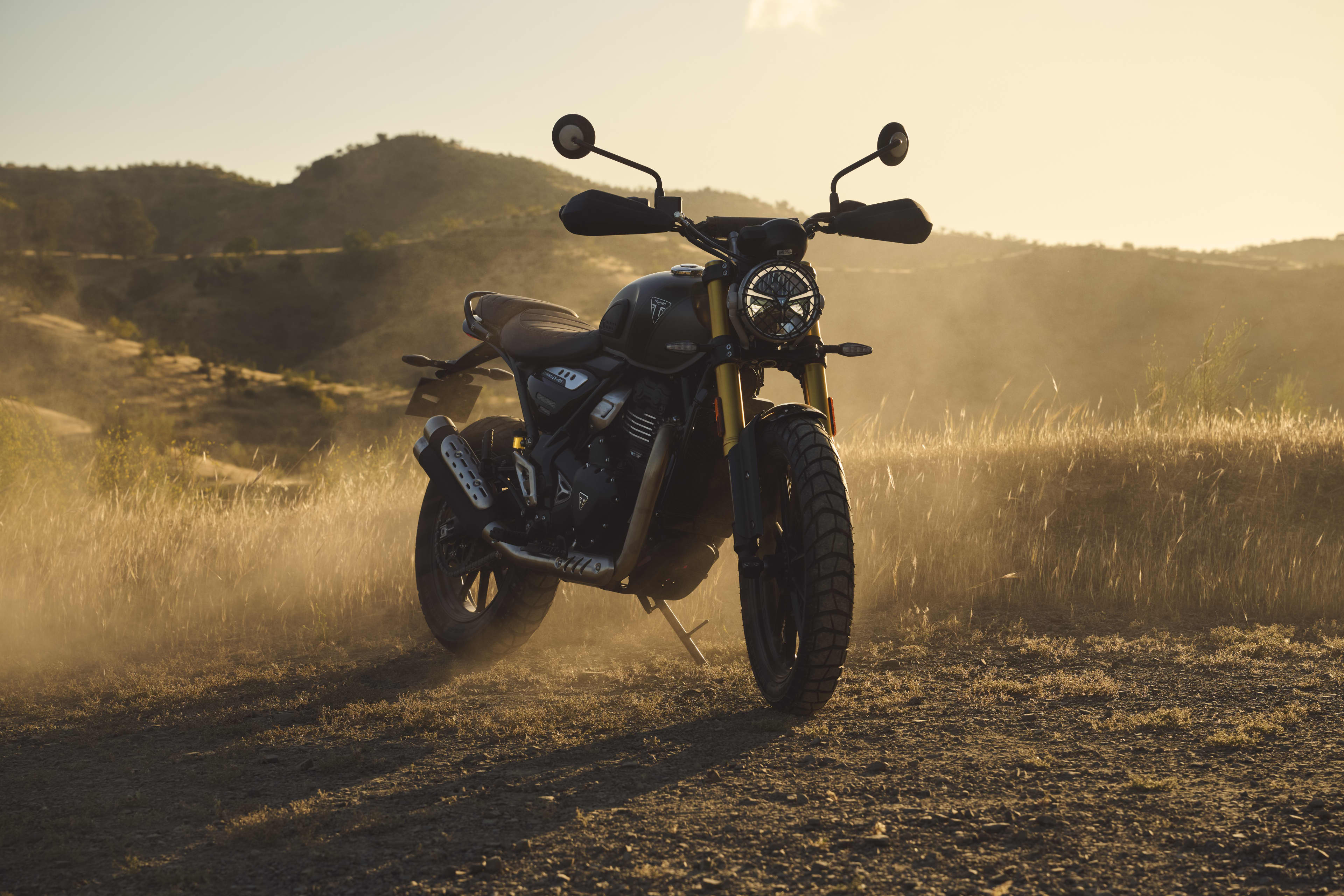 Vue de face du Scrambler 400 X en Matt Pewter Grey et Matt Khaki Green avec un décor poussiéreux tout-terrain.