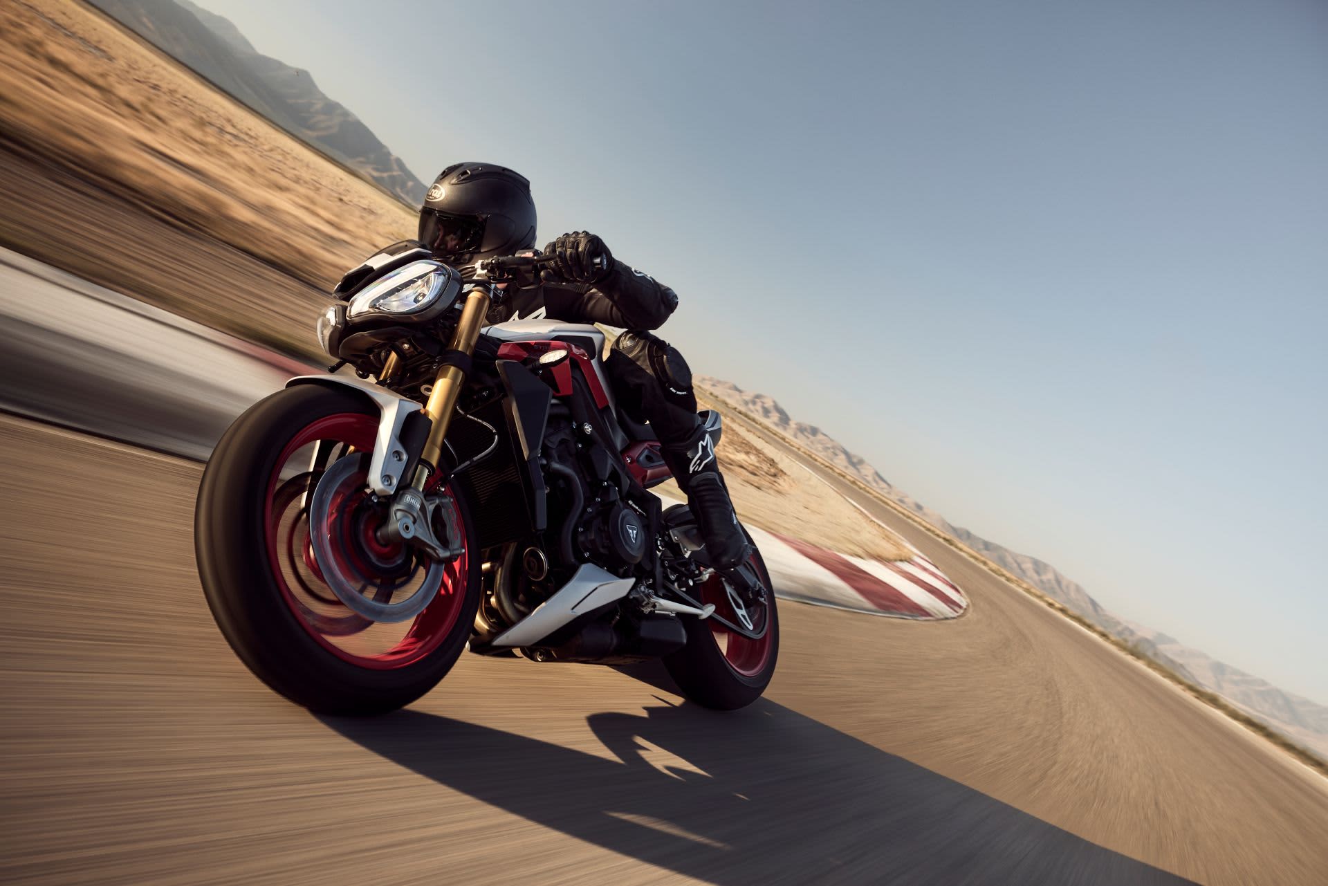 A motorcyclist in black leathers leaning heavily into a high-speed turn on a Triumph Street Triple 765 RX sportbike, showcasing its distinctive red wheels.