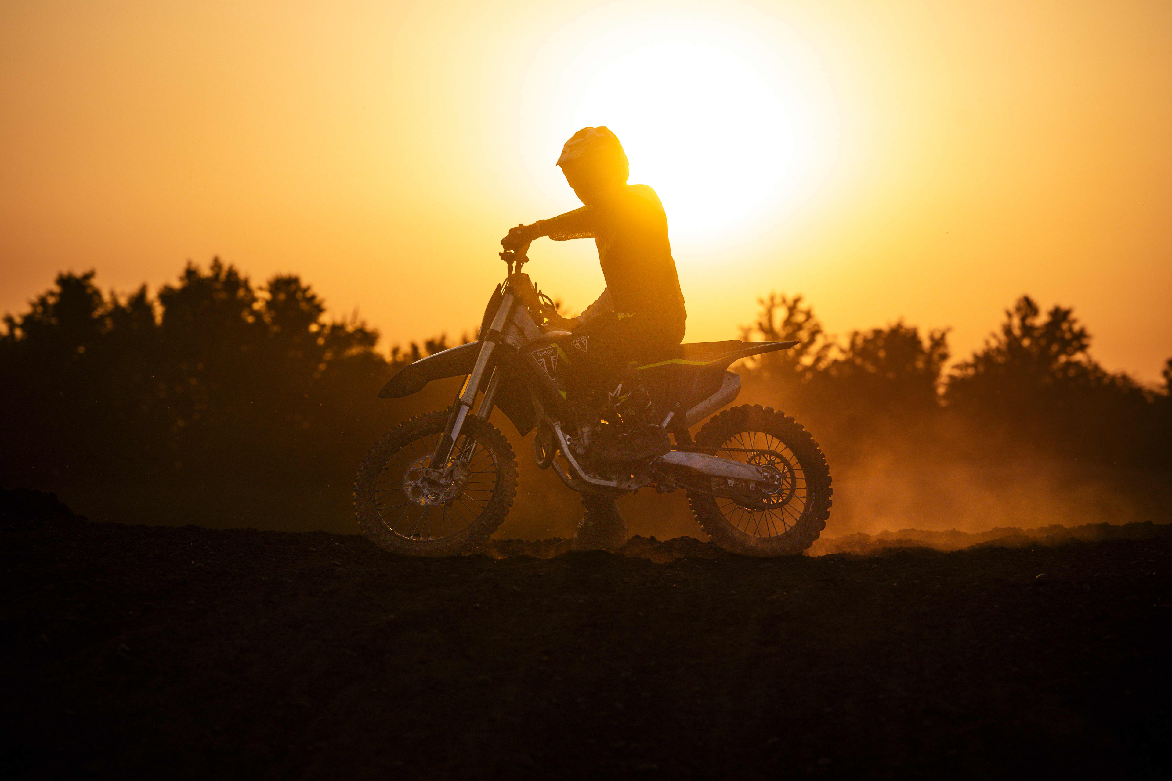 Dramatic sunset photo showing the dark silhouette of a rider sitting on a Triumph 250-X motocross bike against a glowing orange and yellow sky with tree outlines in the background.