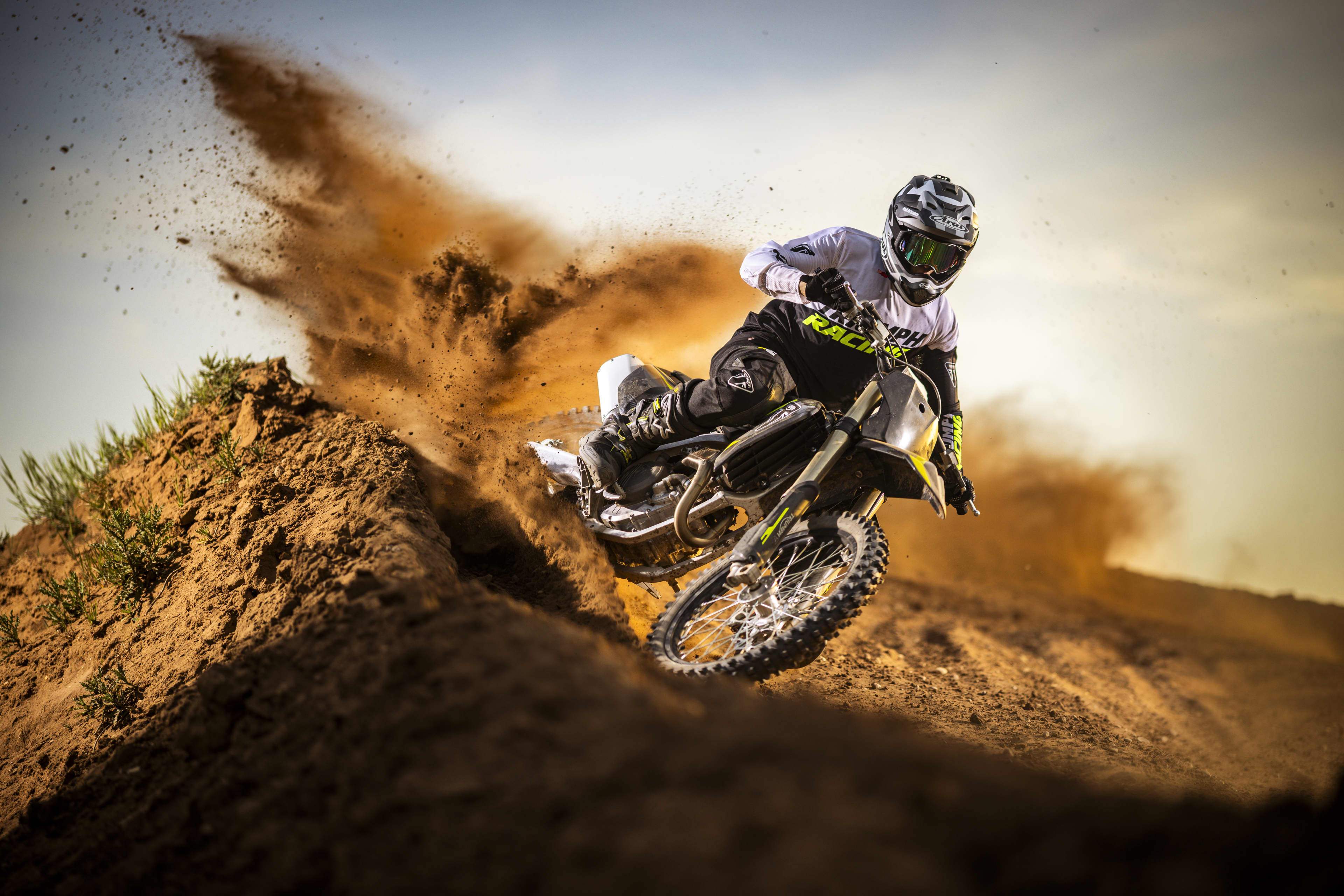 A motocross rider in full gear carving through a dirt berm, throwing up a large cloud of dust riding a Triumph 250-X.