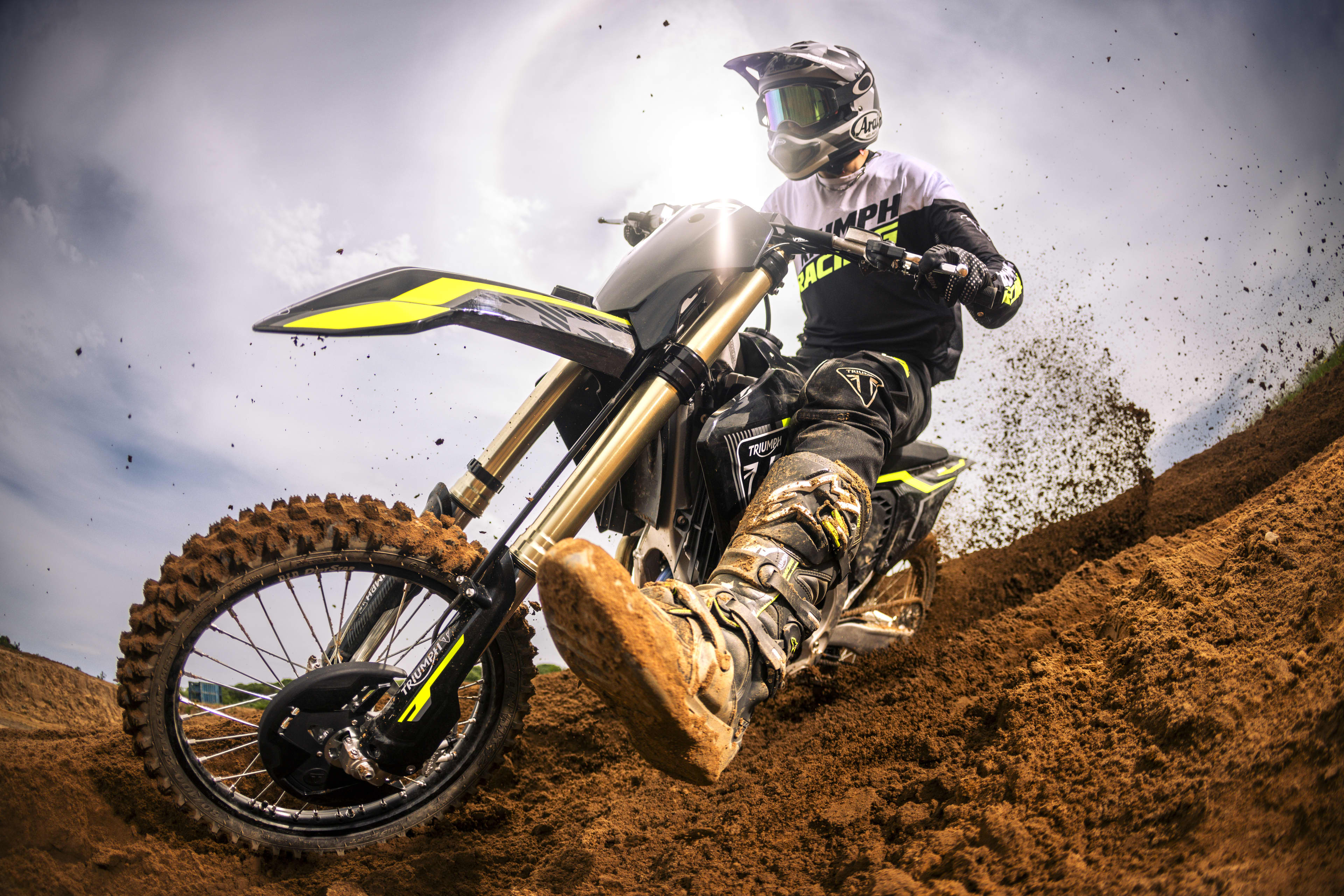 Action-packed low-angle shot of a helmeted rider in Triumph Racing gear riding a Triumph TF 250-X dirt bike through a muddy berm, kicking up dirt.