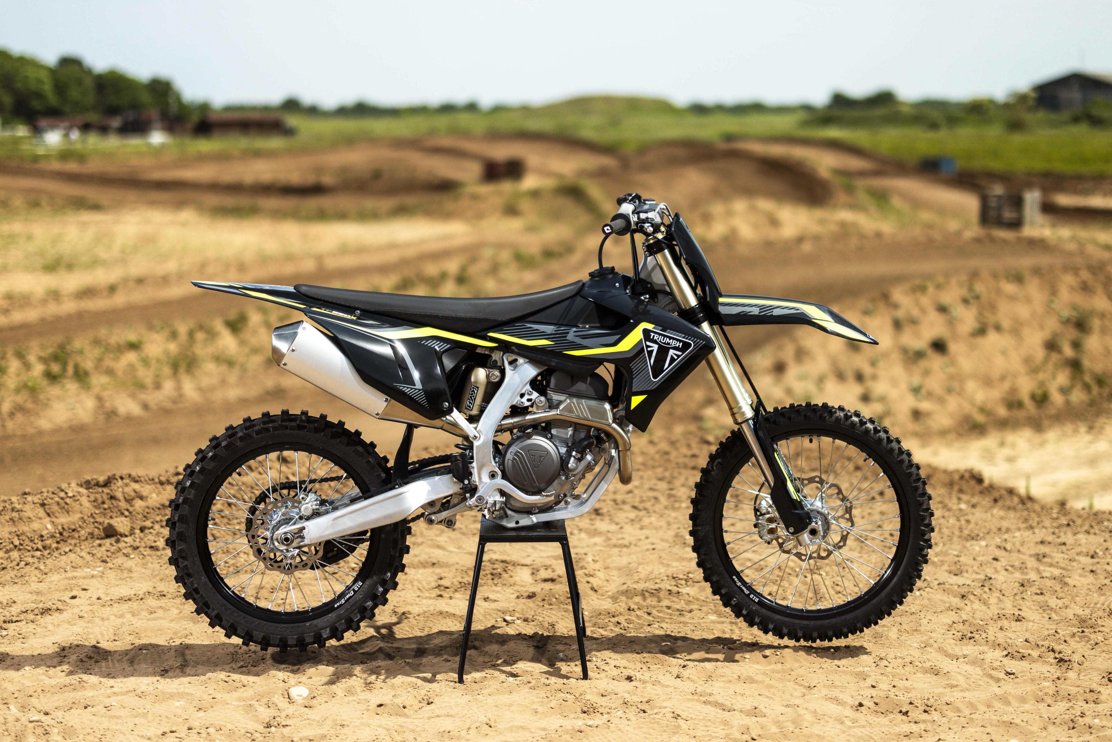 A black and yellow Triumph 250-X dirt bike parked on a central stand at an off-road track.