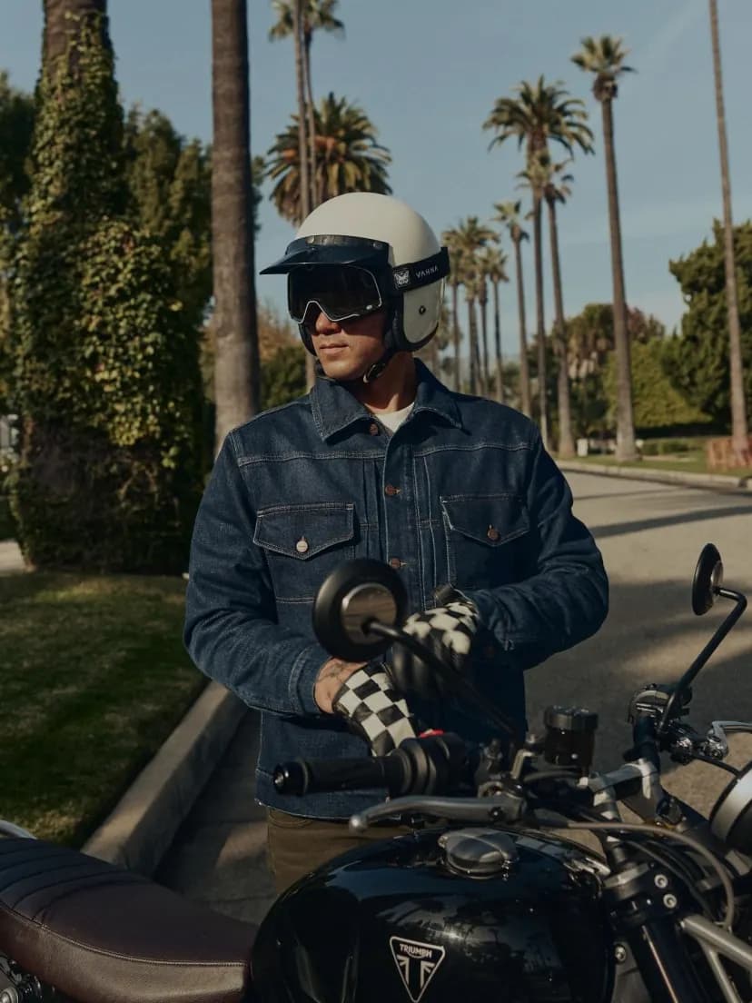 A helmeted rider wearing a dark blue jacket sits on a motorcycle beside a sunlit road lined with palm trees.
