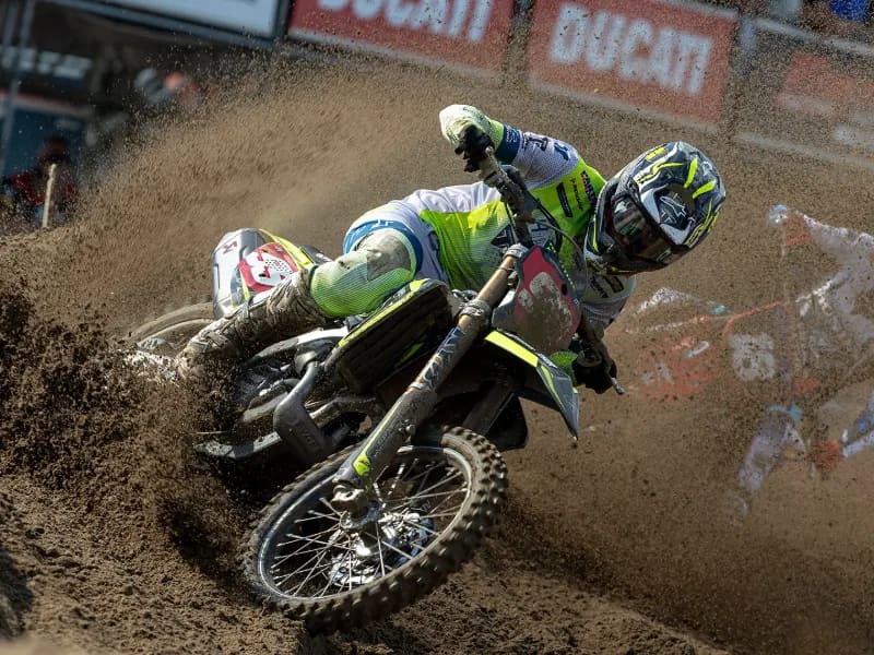 Motocross riders in fluorescent yellow and black racing gear lean their motorcycles through a dirt-track corner.
