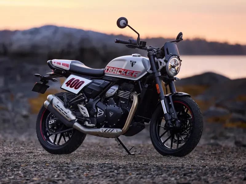 A silver and black Triumph Tracker 400 motorcycle with flat-track styling, side number plates, and "TRACKER" graphics parked on a gravel shore at sunset.