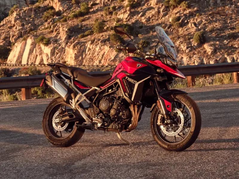 A red and black Triumph Tiger 900 GT adventure motorcycle parked on an asphalt road with rocky cliffs in the background.