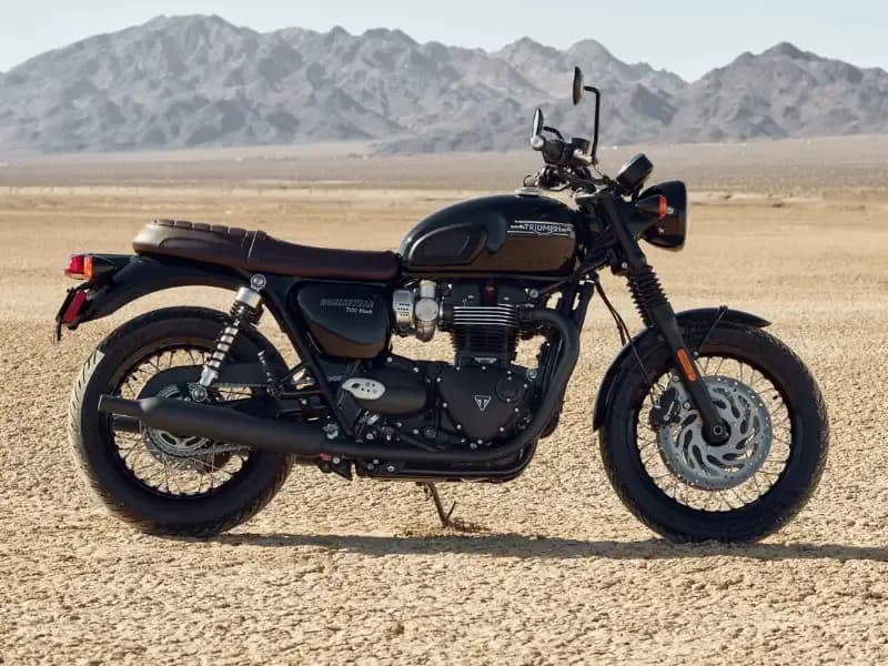 Side profile of a black Triumph Bonneville T120 motorcycle with a brown leather seat parked in a desert landscape.