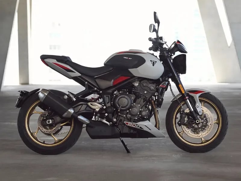 A side profile shot of a white and black Triumph Trident 800 roadster motorcycle with gold wheels, parked inside a modern concrete pavilion featuring large structural pillars.