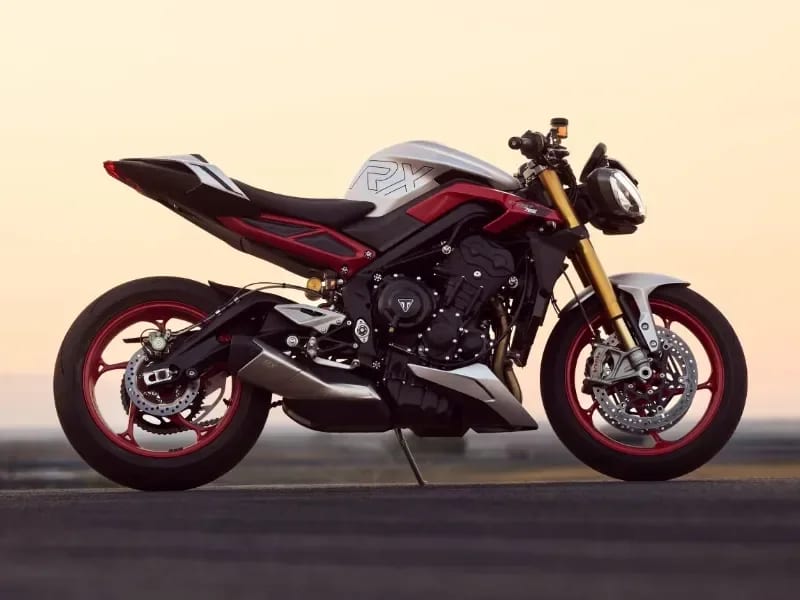 A side profile shot of a silver, red, and black Triumph Street Triple 765 RX naked sport motorcycle with red wheels, parked on an open asphalt track during a hazy golden sunset.
