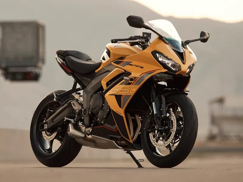A yellow and black Triumph Daytona 660 sports motorcycle parked on a racetrack pit lane, with a race control tower and mountains visible in the blurred background.