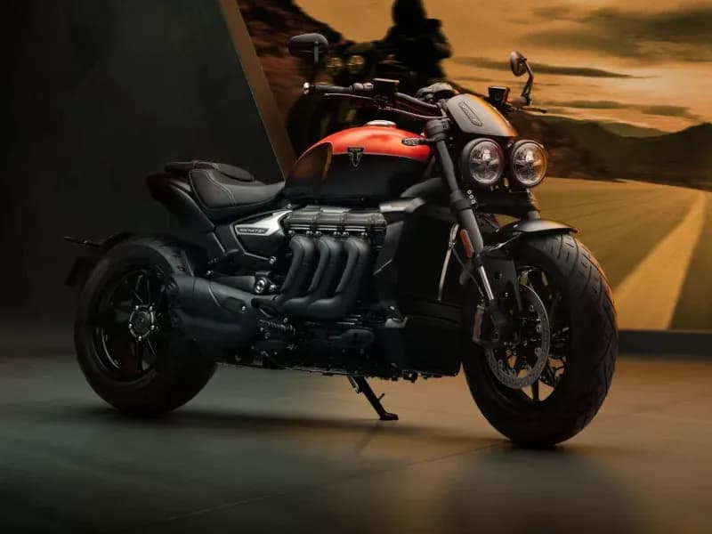 A black and red Triumph Rocket 3 Storm R cruiser motorcycle parked on a showroom floor in front of a scenic desert highway backdrop.
