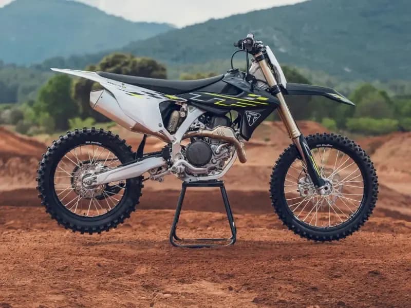 A side profile shot of a Triumph TF 250-X off-road bike resting on a central paddock stand at an outdoor dirt track with hills in the background.