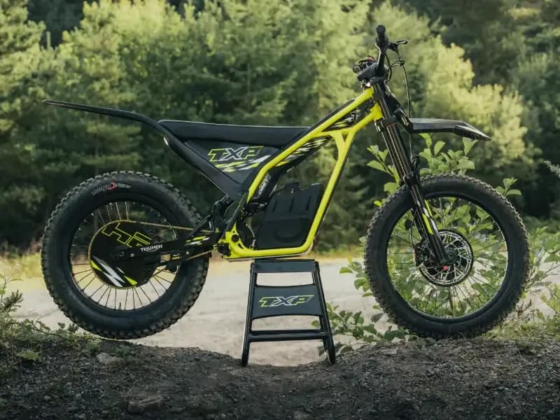 A side profile shot of a neon yellow and black Triumph TXP electric off-road bike resting on a paddock stand on a dirt path with a lush forest background.