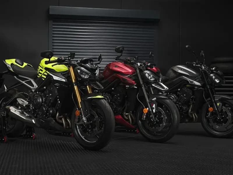 Three modern Triumph Street Triple roadster motorcycles—one in vibrant yellow, one in deep red, and one in sleek grey—parked side-by-side inside a dark, professional garage setting with a black roller door in the background.
