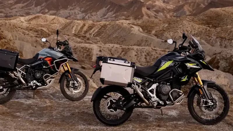 Two Triumph adventure motorcycles—a Tiger 900 and a Tiger 1200—parked on a rocky desert landscape at sunset with mountains in the background.