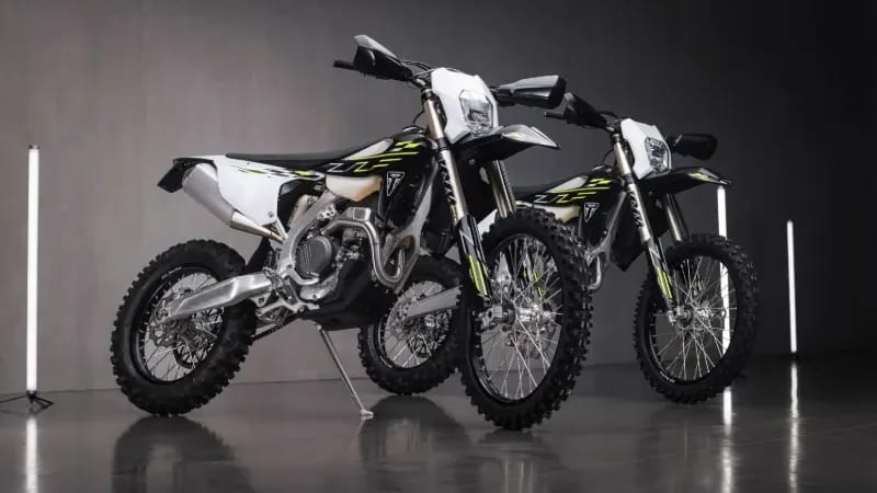 Two Triumph enduro motorbikes parked on a glossy dark floor in a studio setting with vertical tube lights.