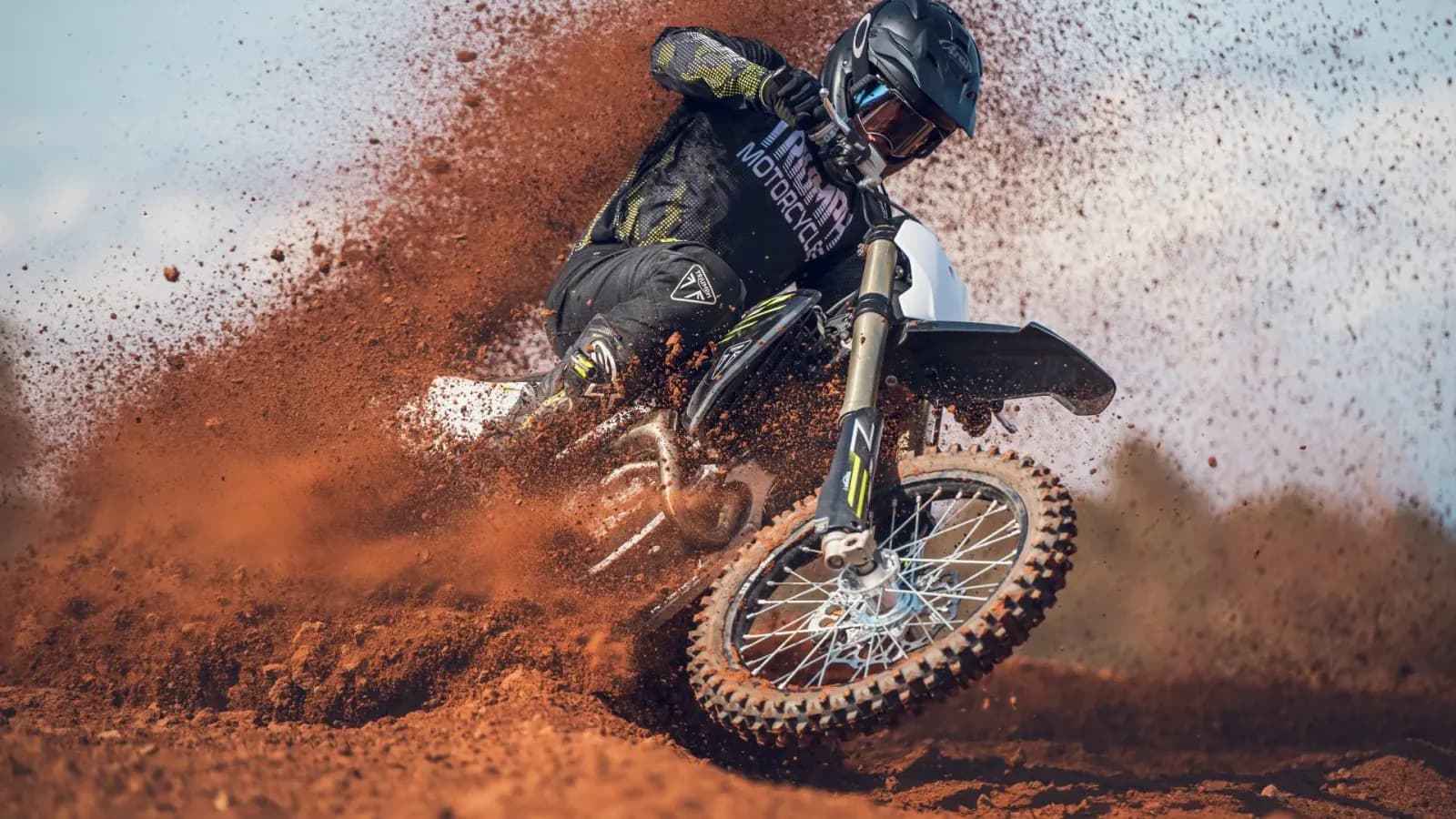 A motocross rider in full gear tightly corners a Triumph TF 450-X dirt bike, aggressively blasting thick red dirt and roost into the air.