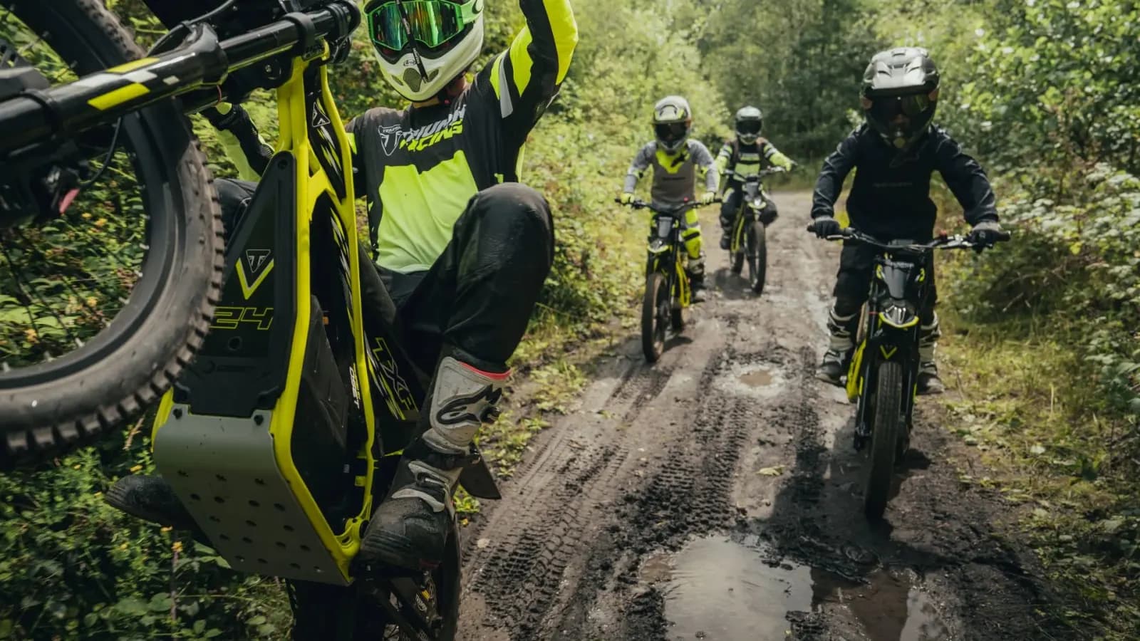 A group of young riders navigating a muddy, tree-lined trail on neon yellow Triumph TXP electric dirt bikes, with the lead rider performing a wheelie.