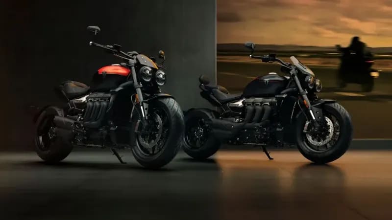 Two Triumph Rocket 3 Storm motorcycles—the R model with a red tank accent and the GT model in all-black—parked side-by-side in a dimly lit studio showroom.