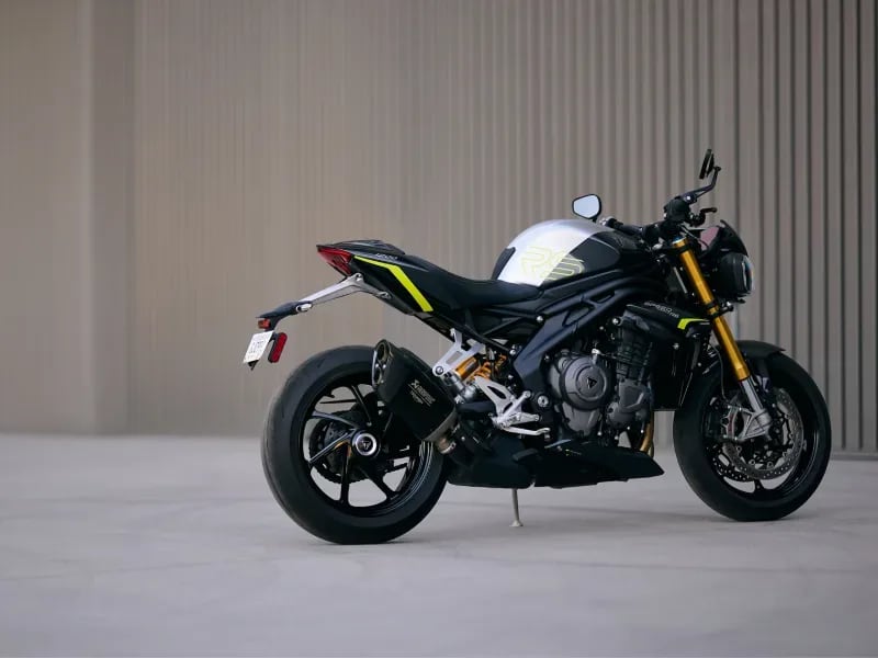 Side profile of a black and silver Triumph Speed Triple RS motorcycle with yellow accents parked outdoors.