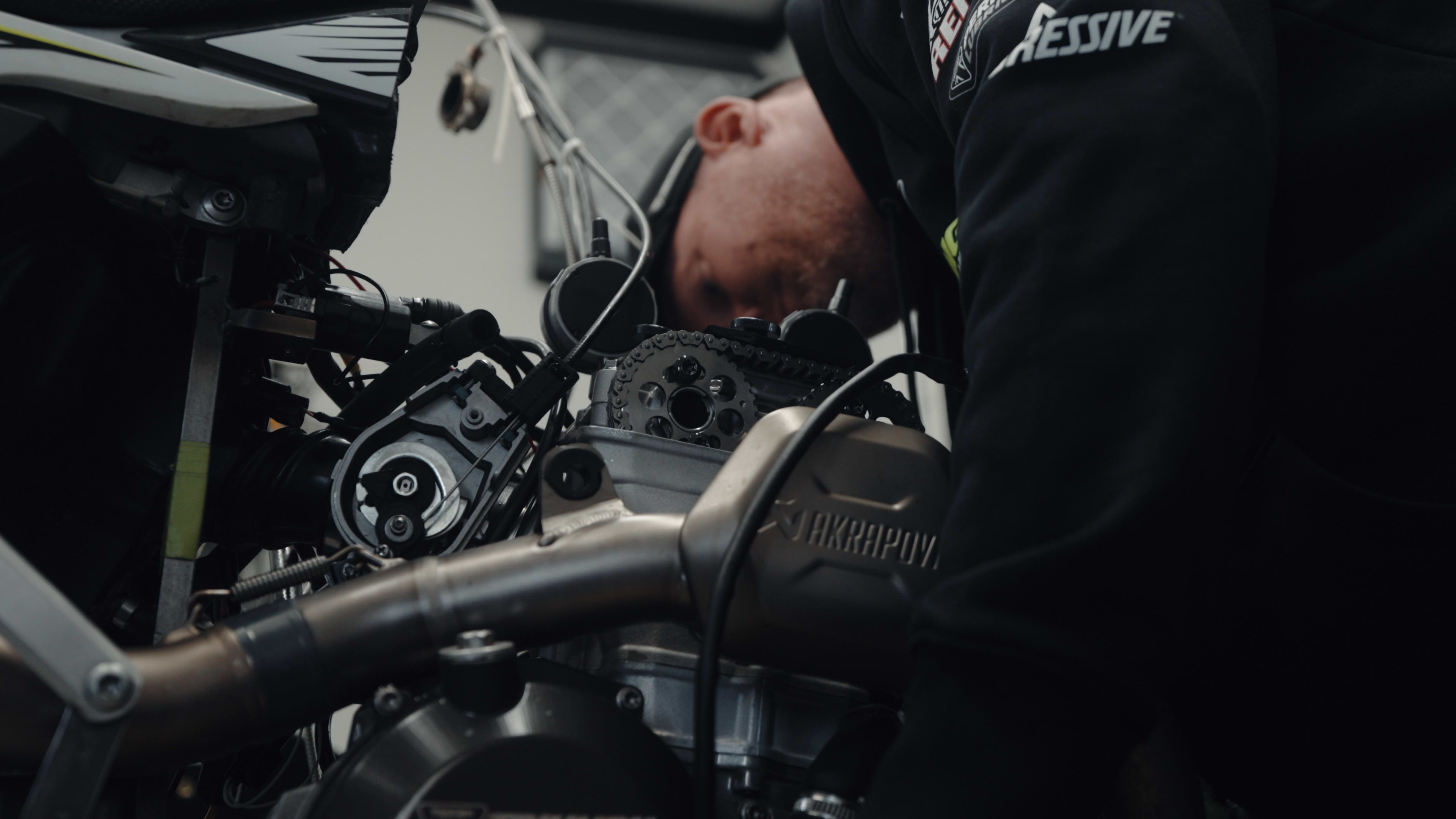 Medium close-up shot of a race mechanic inspecting the cylinder head, exposed timing chain, and camshaft sprocket of a Triumph TF 250-X engine featuring an integrated Akrapovič exhaust system.
