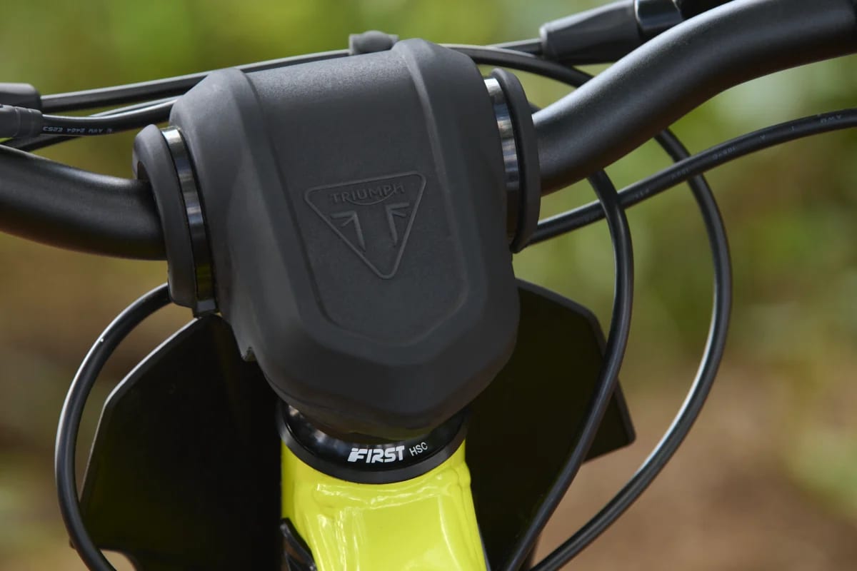 Macro shot showcasing the matte black protective handlebar pad with an embossed Triumph logo, mounted above the headset bearing and neon green neck frame of a Triumph TXP-20 youth electric motorcycle.