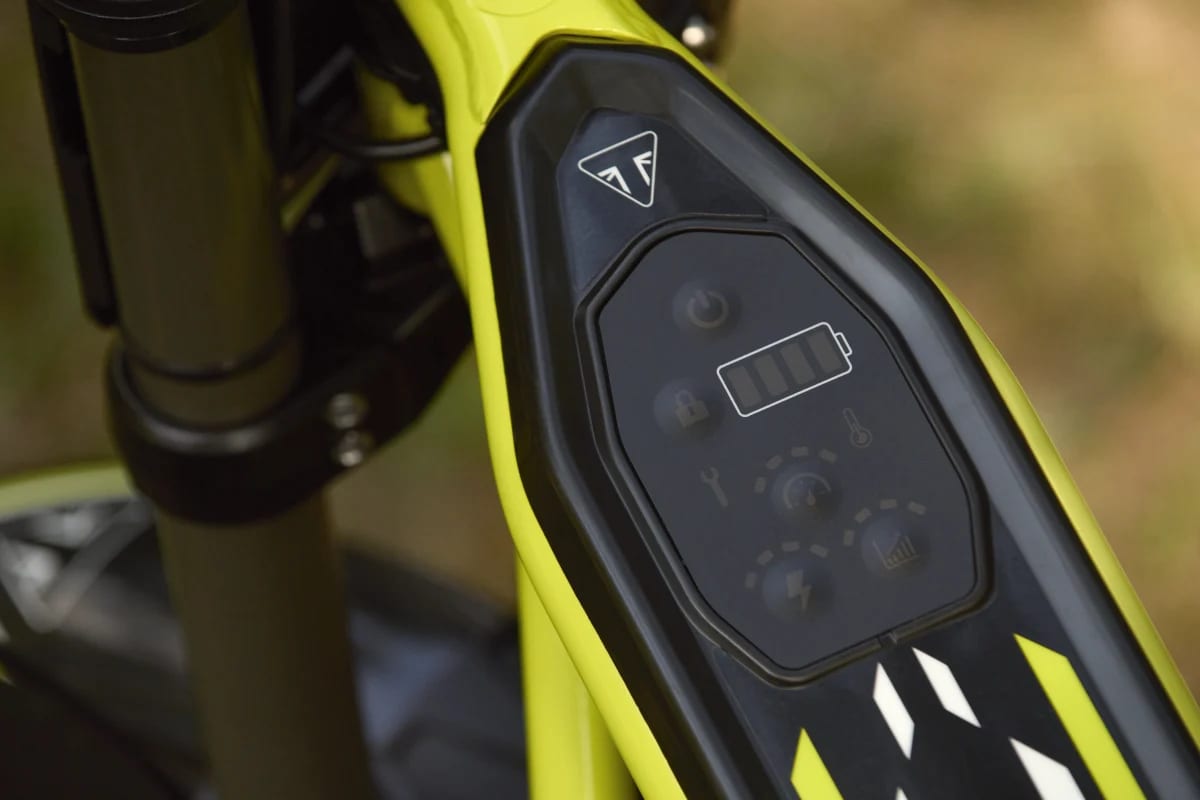 Frame-mounted digital interface panel with power button, battery charge indicator, speed controls, and Triumph badge on a TXP-20 youth electric motorcycle.