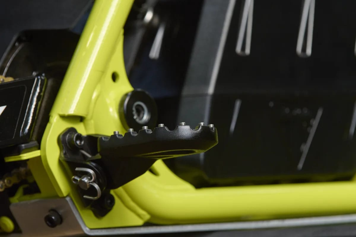 Macro shot showing a high-grip black serrated metal footpeg with retaining pin and spring assembly, mounted to the neon green lower frame and skid plate section of a Triumph TXP-20 youth electric motorcycle.