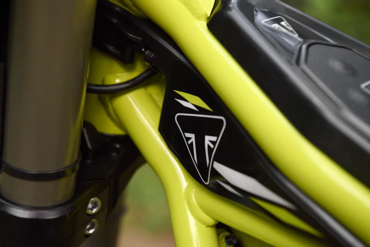 Extreme close-up shot highlighting the black triangular Triumph badge with white and neon yellow accents on the upper tubing of the neon green frame, positioned near the front suspension and top cover of a TXP-20 youth electric motorcycle.