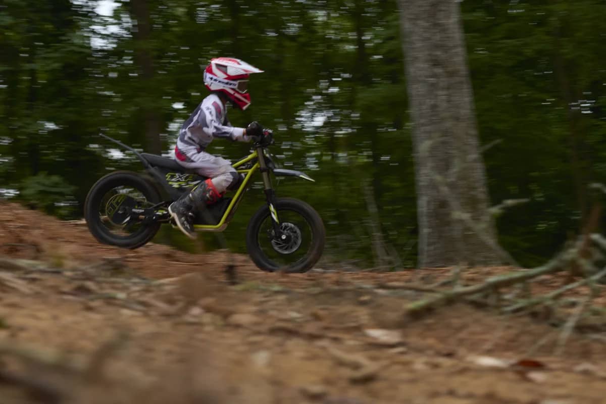 Side-profile motion shot capturing a young rider in full motocross gear taking a jump on a Triumph TXP-20 youth electric motorcycle, surrounded by woodland trees with panning blur.