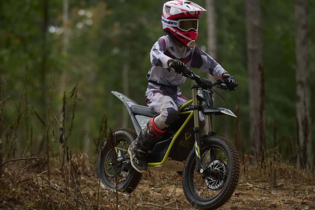 Side profile action view of a young rider wearing full gear and helmet maneuvering a Triumph TXP-20 electric dirt bike over a log along a forest trail.