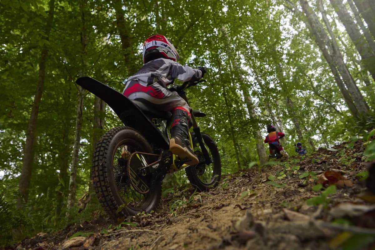 Low-angle rear action shot following a youth rider in protective riding gear as they navigate a Triumph TXP-20 electric dirt bike up a dirt slope through a dense, sunlit forest.