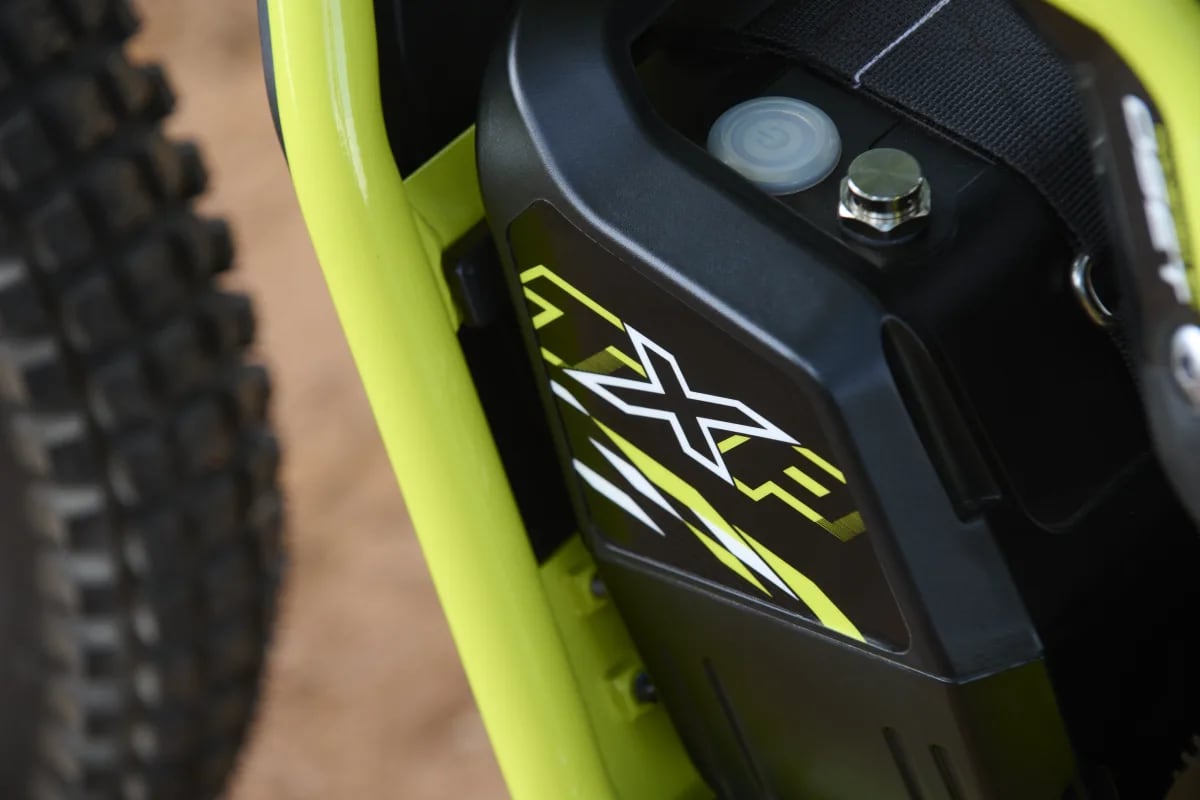 Close-up view of the lithium-ion battery pack featuring TXP graphic decals, power button, and magnetic safety lanyard mount set within the neon green tubular frame of a Triumph TXP-20 youth electric motorcycle.