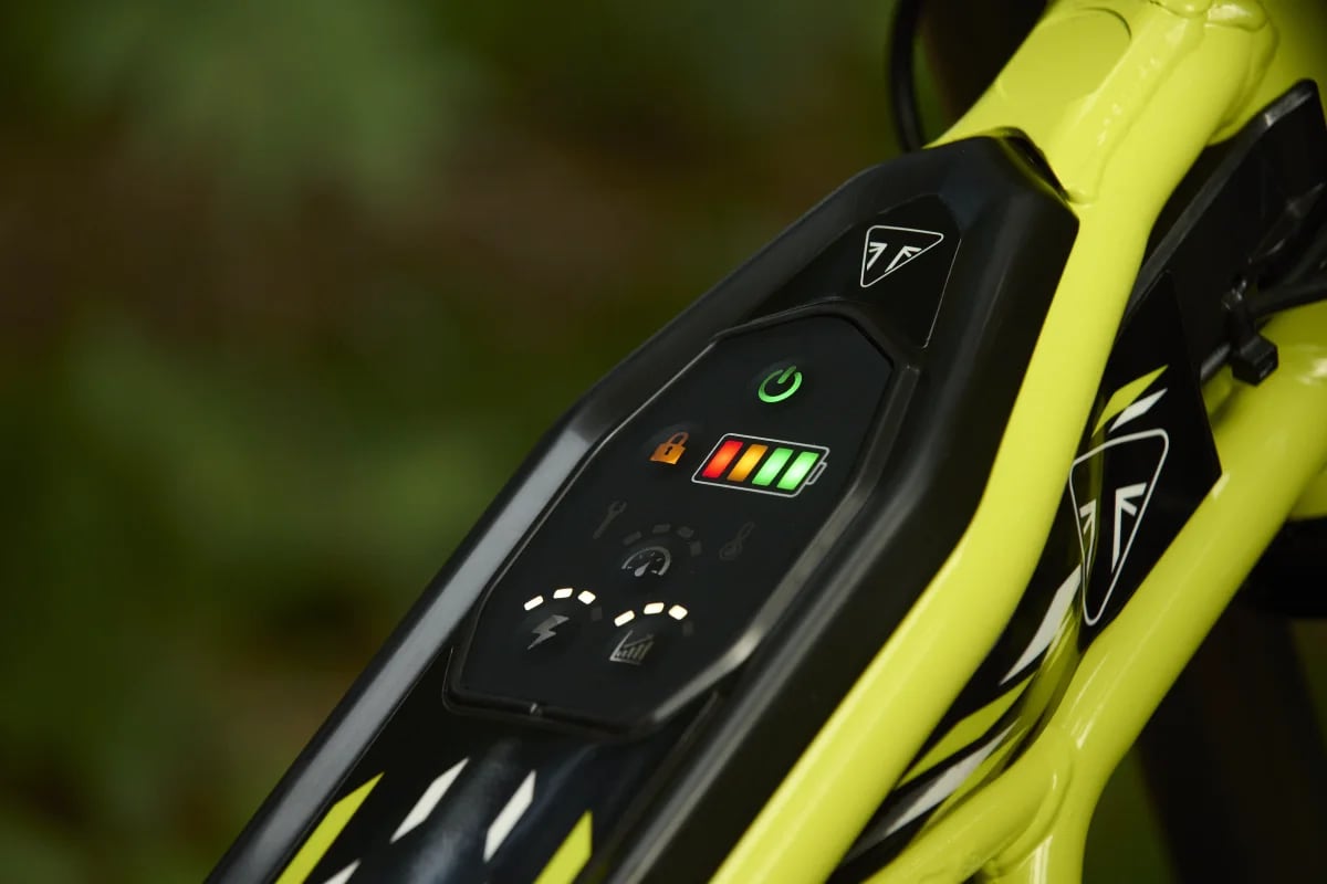 Macro shot focusing on the illuminated digital display module mounted on the top frame tube of a Triumph TXP-24 electric motorcycle, displaying a green power icon, colorful LED battery life gauge, and power mode status indicators framed by neon yellow tubes and Triumph branding.