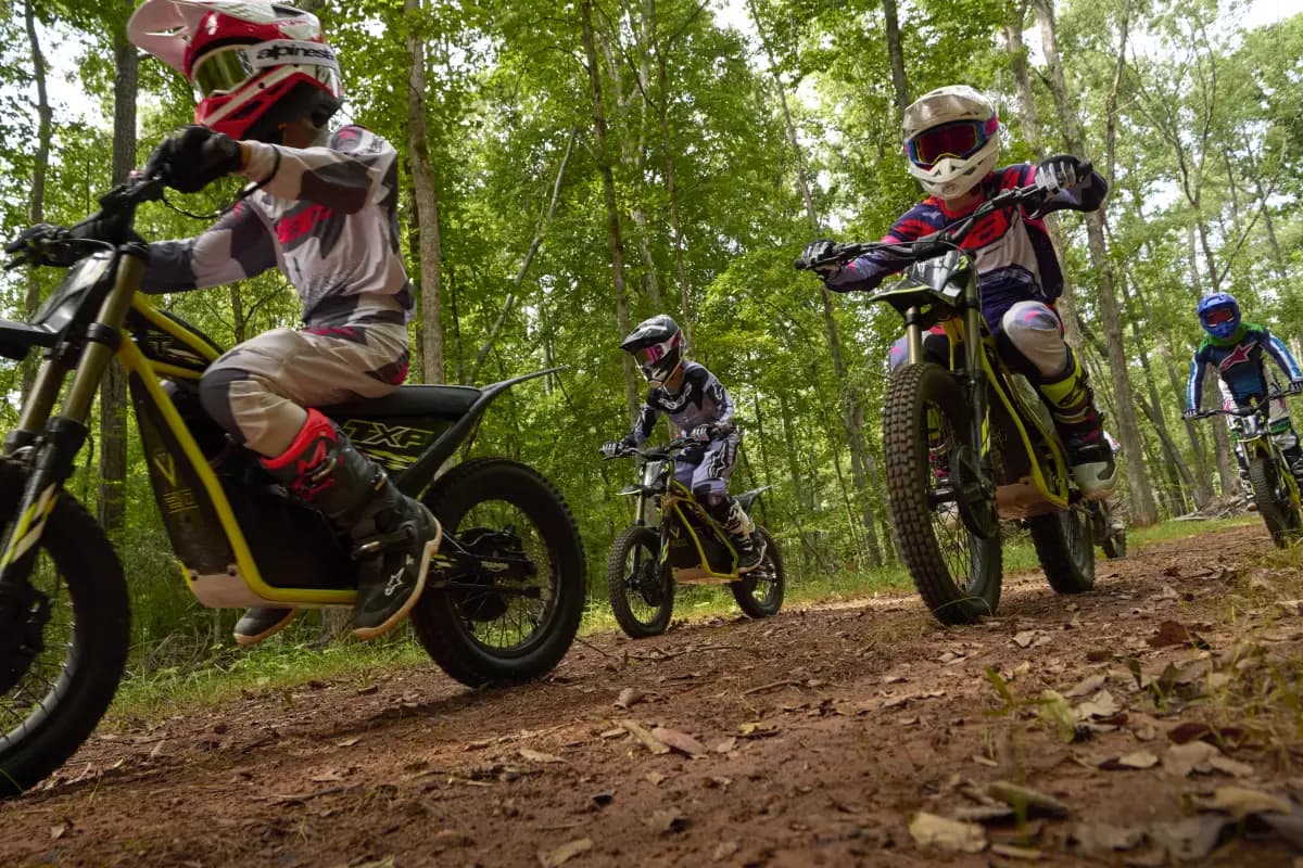 Low-angle dynamic group action shot of several young riders in full protective helmets and motocross gear riding Triumph TXP-20 electric dirt bikes along a dirt forest trail under a bright canopy of trees.