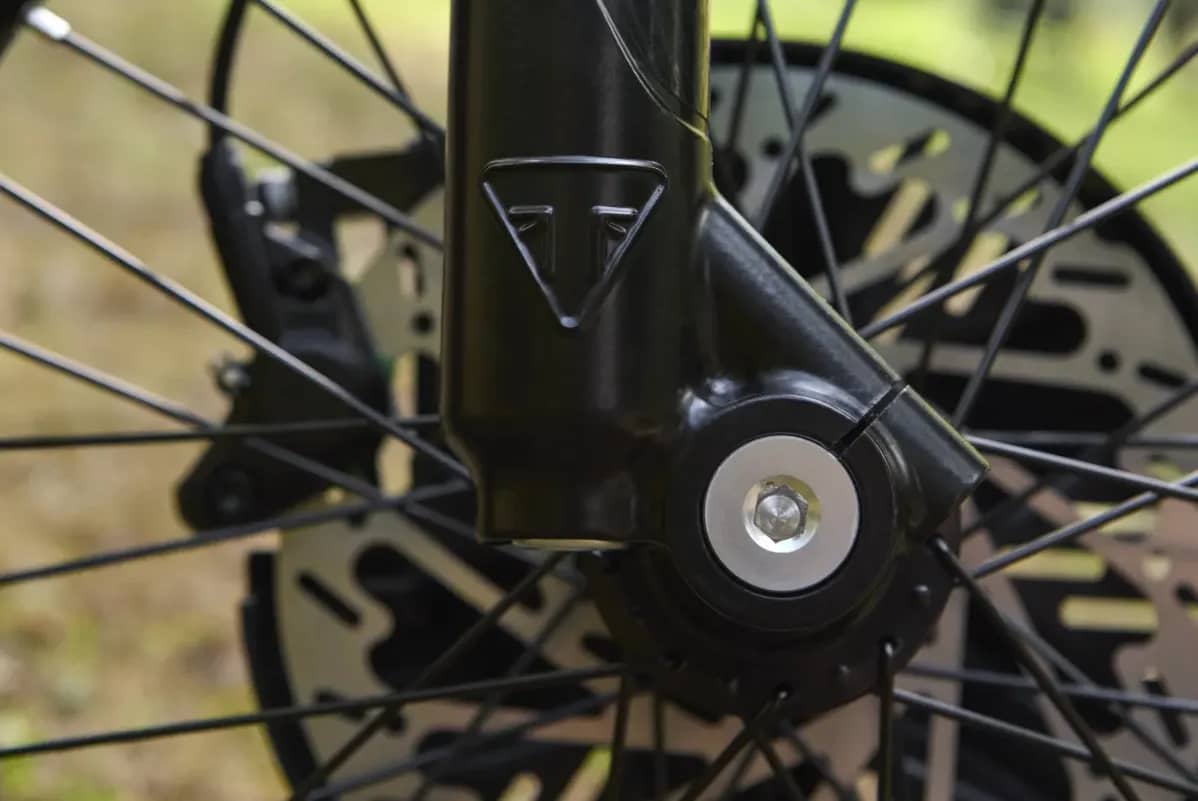 Macro detail shot showing an embossed Triumph logo on the black lower fork leg, highlighting the front axle bolt, spoke pattern, brake caliper, and ventilated disc brake rotor on a TXP-20 youth electric motorcycle.