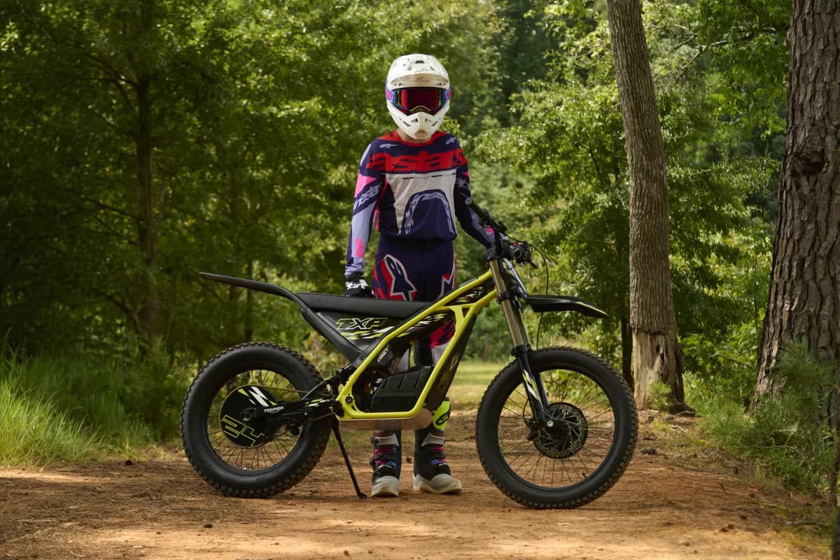 Full-length outdoor shot of a young rider fully outfitted in a white helmet and blue, white, and pink gear standing next to a side-profile Triumph TXP-24 electric motorcycle parked on a kickstand along a dirt forest path.