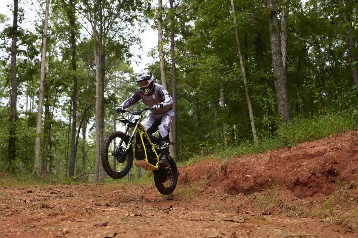 Dynamic action shot showing a young rider fully suited in grey riding gear and a helmet catching air mid-jump on a Triumph TXP-24 electric motorcycle over a red dirt mound in a wooded trail setting.