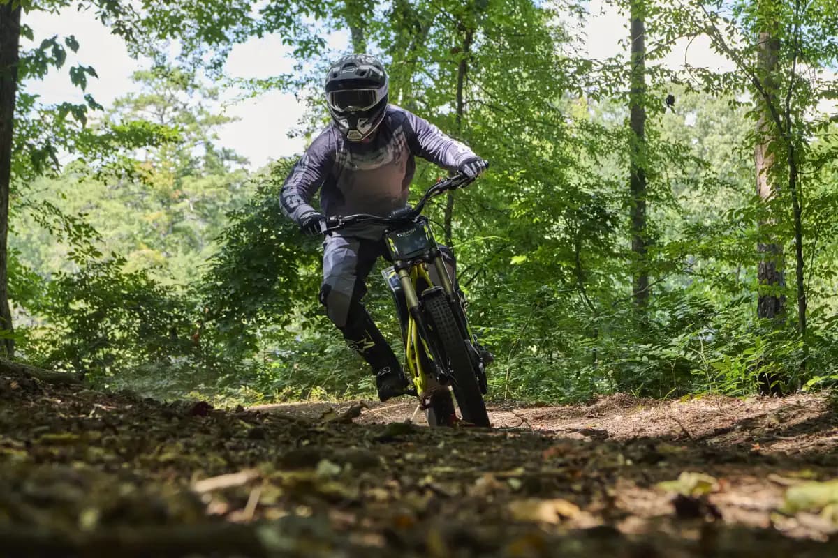 Front-facing action shot showing a rider in full grey motocross gear, helmet, and goggles leaning into a turn on a Triumph TXP-24 electric motorcycle along a shaded dirt path in the woods.