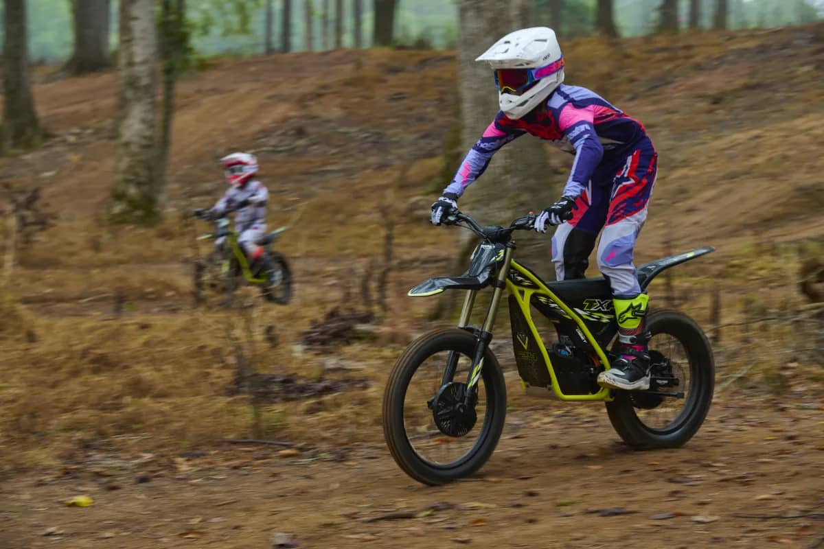 Side profile action shot featuring a young rider in a white helmet, goggles, and pink and purple riding suit operating a Triumph TXP-24 electric motorcycle on a dirt track, with a second youth rider following in the background amid woodland terrain.