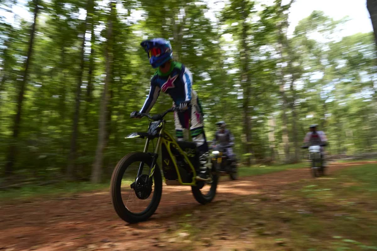 Dynamic motion blur tracking shot capturing a young rider standing on the footpegs of a Triumph TXP-24 electric motorcycle while speeding down a dirt trail in a dense forest, with additional riders following close behind.