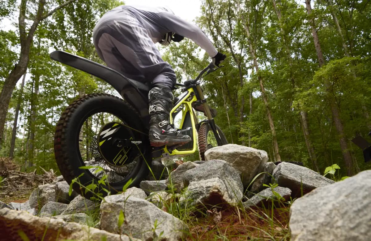 Low-angle dynamic rear action shot showing a young rider in full riding gear and boots standing on the footpegs to balance a Triumph TXP-24 electric motorcycle through a technical rock obstacle in a forest setting.