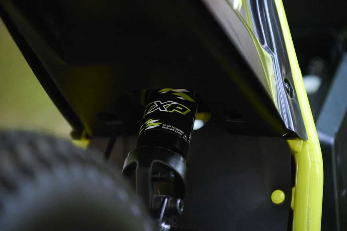 Macro detail shot showing the rear mono-shock absorber with TXP-branded graphics positioned beneath the black rear fender and framed by the neon green chassis structure of a Triumph TXP-24 youth electric motorcycle.