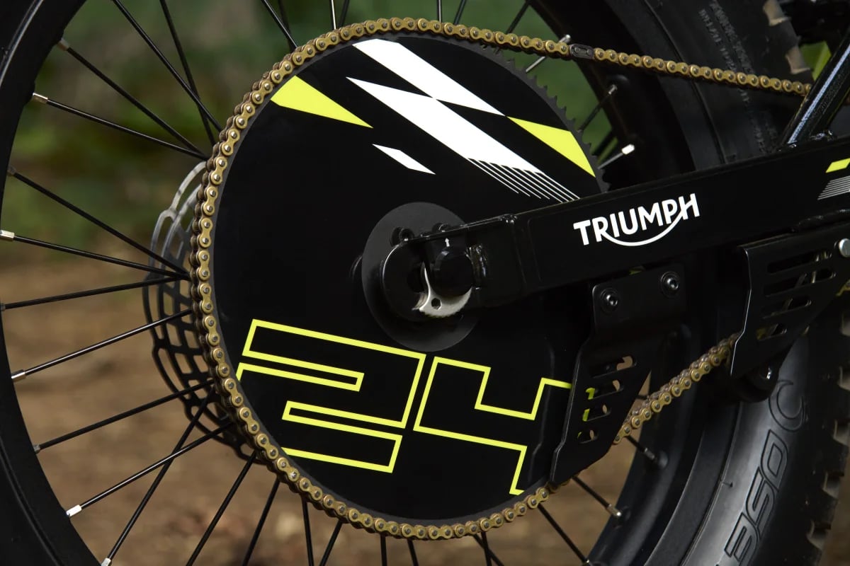 Macro shot highlighting the gold drive chain wrapped around a rear sprocket with a black graphic sprocket guard featuring bold neon yellow '24' lettering and white accents, attached to the black Triumph-branded swingarm of a TXP-24 youth electric motorcycle.