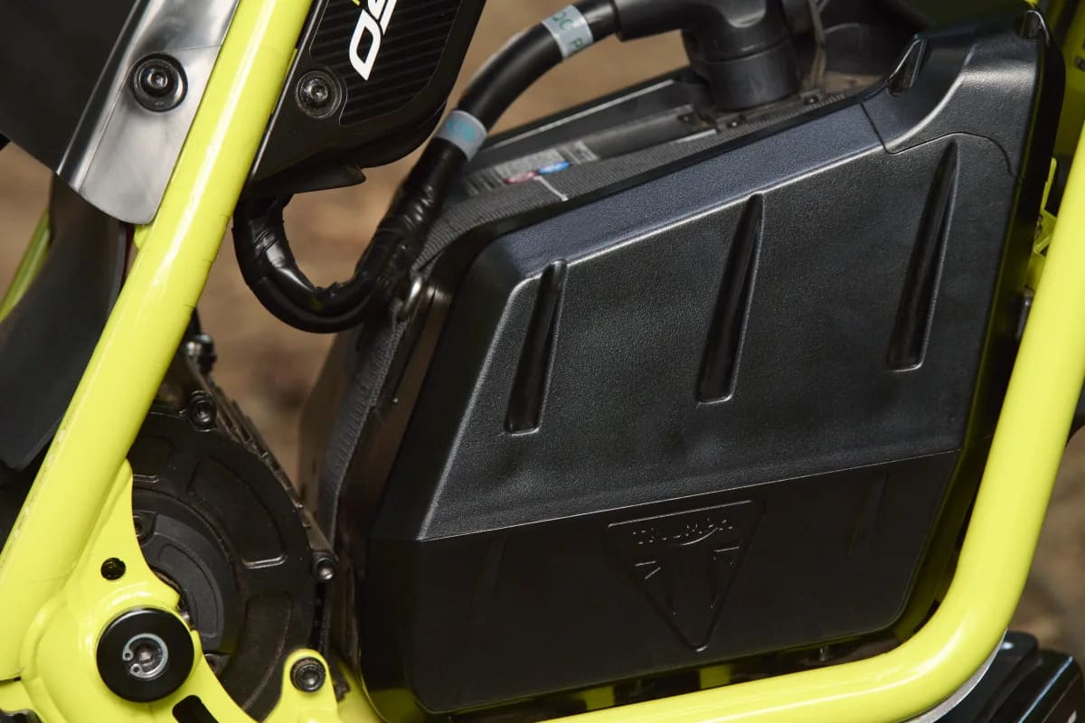 Extreme close-up showcasing the black molded battery pack casing with an embossed Triumph logo nestled within the neon green perimeter frame of a TXP-24 youth electric motorcycle.