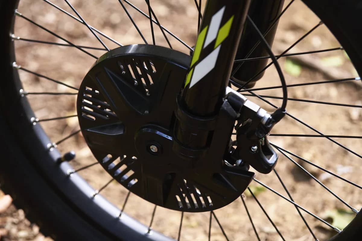 Macro shot showcasing the front wheel assembly of a Triumph TXP-24 electric motorcycle, featuring a black vented front brake disc guard, hydraulic brake caliper, spoked rim, and lower fork leg with neon graphics.