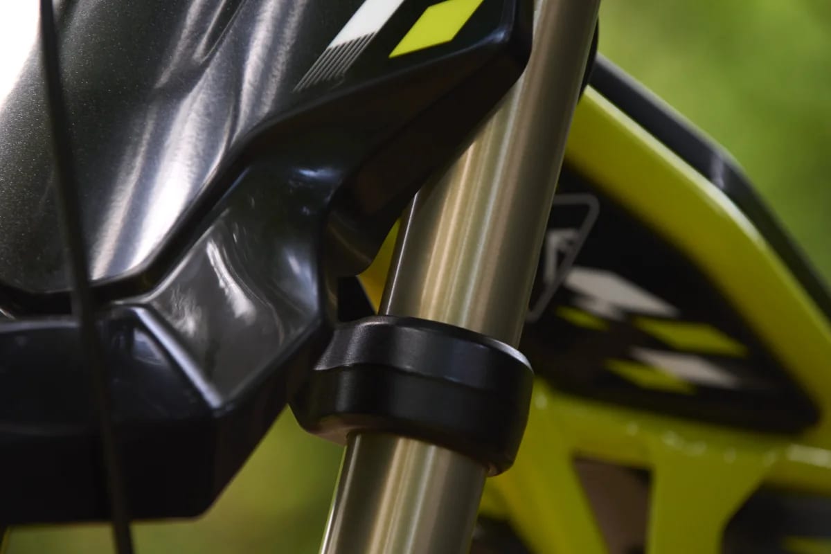 Macro detail shot showing the gold front suspension fork tube where it clamps beneath the black front fender and number plate mount on a Triumph TXP-24 youth electric motorcycle.