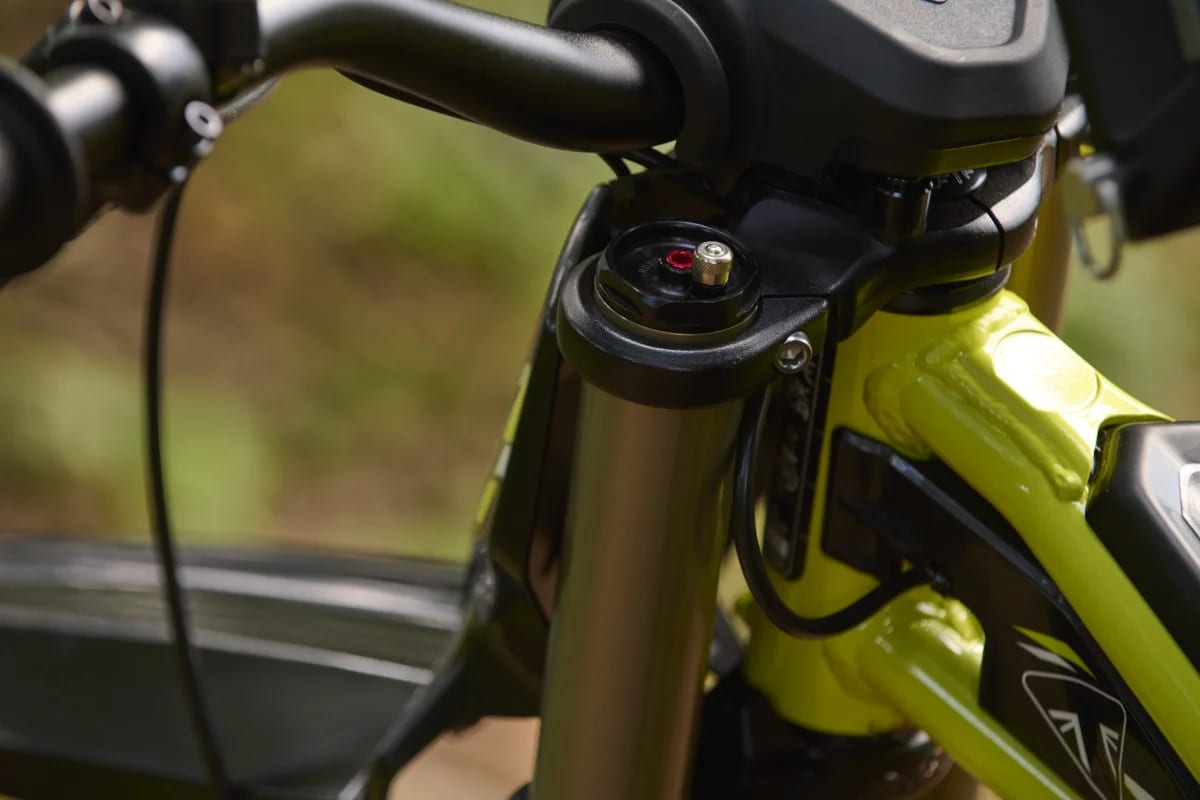 Macro shot highlighting the top fork cap assembly with red suspension adjustment clickers and air valve, clamped into the black upper triple clamp near the handlebars and neon green frame head tube of a Triumph TXP-24 youth electric motorcycle.